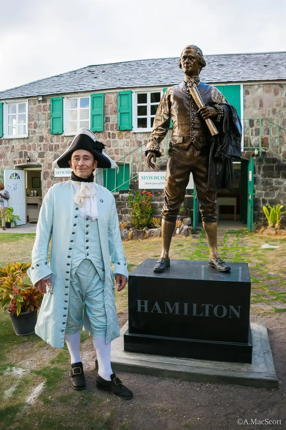 Scott in a light blue colonial coat beside the Alexander Hamilton statue on Nevis